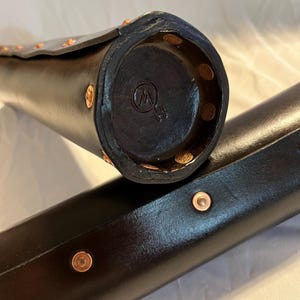 Puede incluir: Primer plano de dos estuches cil&iacute;ndricos de cuero negro con remaches de cobre. Un estuche est&aacute; abierto, revelando un extremo circular con un logotipo estampado. Los estuches parecen hechos a mano y tienen un acabado pulido. Los estuches probablemente est&aacute;n dise&ntilde;ados para guardar un objeto largo y delgado.