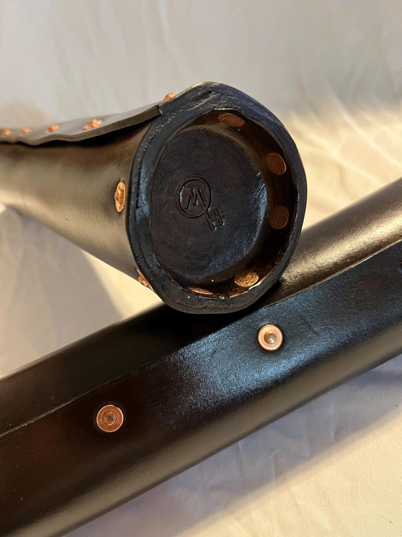 Puede incluir: Primer plano de dos estuches cil&iacute;ndricos de cuero negro con remaches de cobre. Un estuche est&aacute; abierto, revelando un extremo circular con un logotipo estampado. Los estuches parecen hechos a mano y tienen un acabado pulido. Los estuches probablemente est&aacute;n dise&ntilde;ados para guardar un objeto largo y delgado.