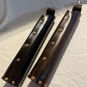 Puede incluir: Se muestran dos brazales de cuero, uno negro y otro marr&oacute;n. Cada brazalete cil&iacute;ndrico tiene remaches de cobre y un lazo en la parte superior. Estos brazales son probablemente para tiro con arco o recreaciones hist&oacute;ricas.