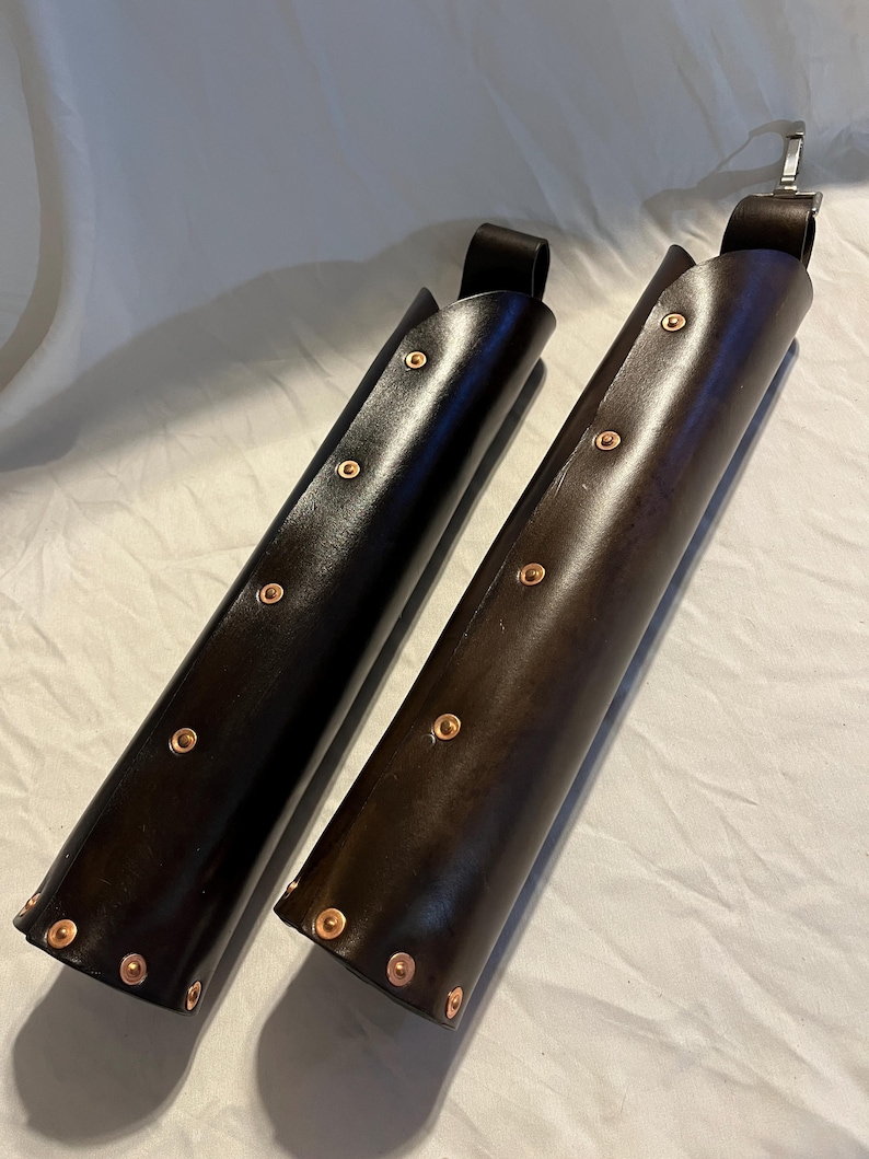 Puede incluir: Se muestran dos brazales de cuero, uno negro y otro marr&oacute;n. Cada brazalete cil&iacute;ndrico tiene remaches de cobre y un lazo en la parte superior. Estos brazales son probablemente para tiro con arco o recreaciones hist&oacute;ricas.