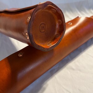 May include: Close-up of two brown leather cases. The cases are cylindrical with a rounded bottom and a flap closure. Copper rivets secure the leather. The bottom of one case has a stamped logo. The leather has a rich, warm tone.
