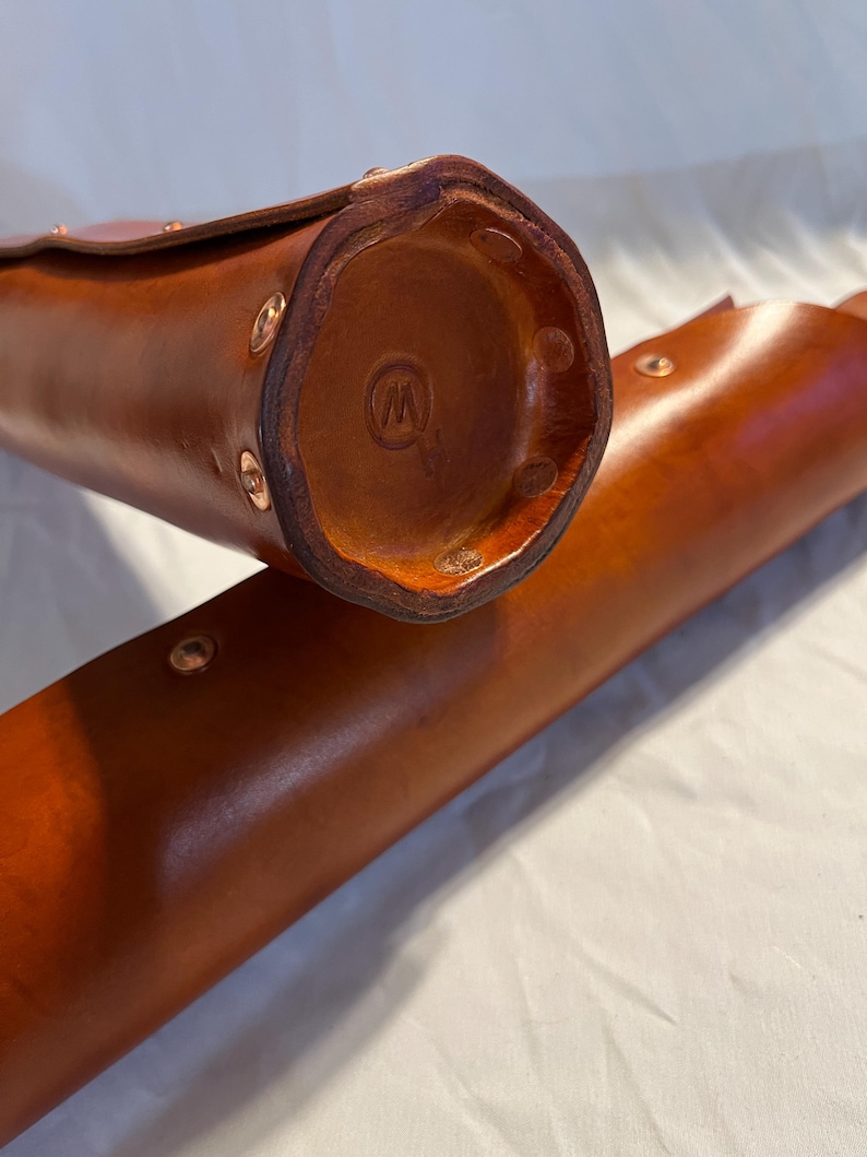 May include: Close-up of two brown leather cases. The cases are cylindrical with a rounded bottom and a flap closure. Copper rivets secure the leather. The bottom of one case has a stamped logo. The leather has a rich, warm tone.