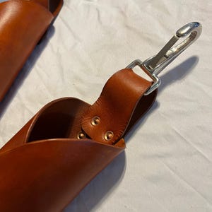 May include: Two brown leather sheaths with a loop handle and a silver-colored metal clasp. The leather is a rich, warm brown color. The sheaths appear to be handmade, with visible stitching and rivets. The background is a white fabric.