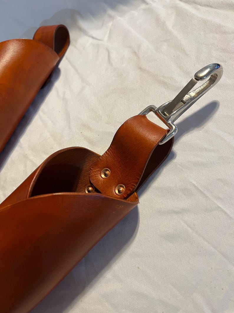 May include: Two brown leather sheaths with a loop handle and a silver-colored metal clasp. The leather is a rich, warm brown color. The sheaths appear to be handmade, with visible stitching and rivets. The background is a white fabric.