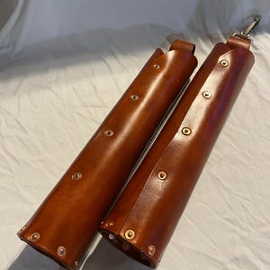 May include: Two brown leather cases with brass rivets and a silver clasp. The cases are cylindrical and appear to be handmade. The leather has a rich, warm tone, and the rivets are evenly spaced along the sides.