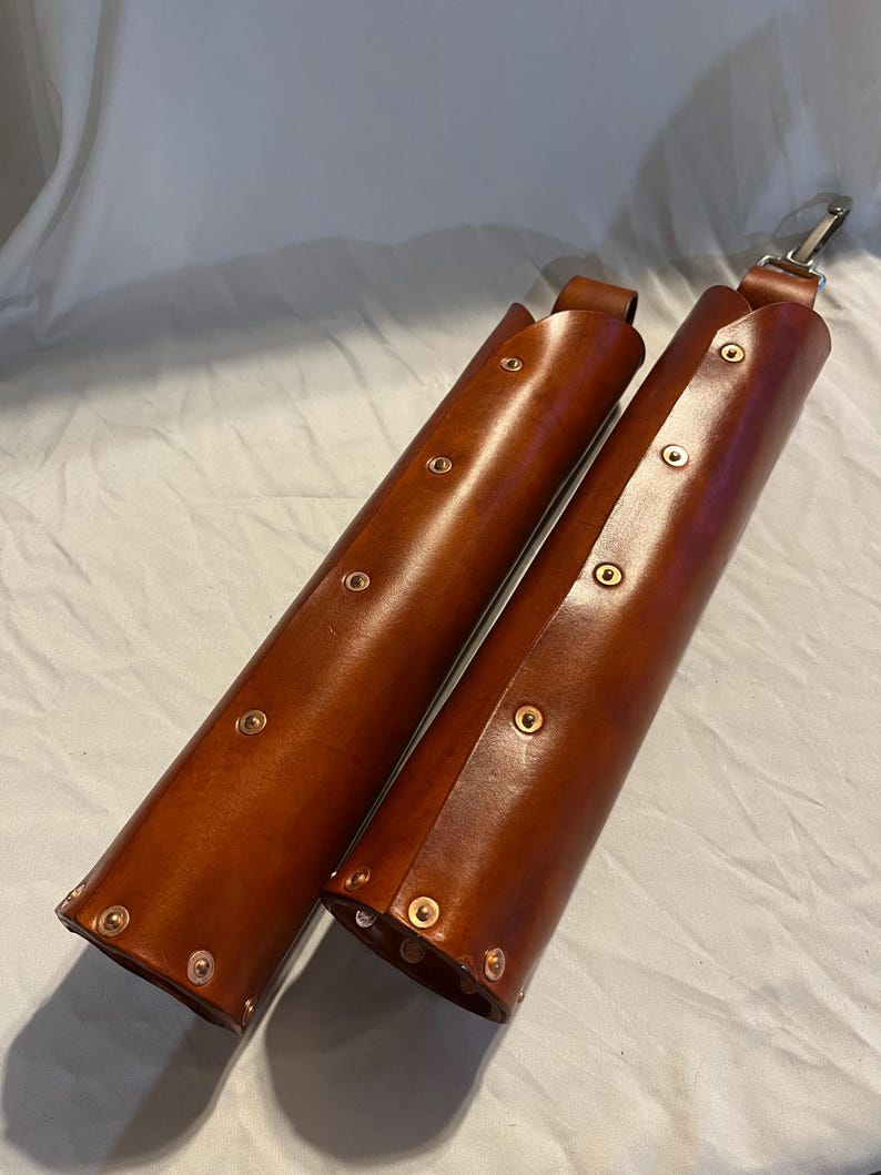 May include: Two brown leather cases with brass rivets and a silver clasp. The cases are cylindrical and appear to be handmade. The leather has a rich, warm tone, and the rivets are evenly spaced along the sides.