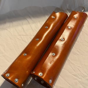 May include: Two brown leather arm guards with silver rivets. The cylindrical guards are open on one side and have a metal clasp at the top. The leather has a glossy finish.