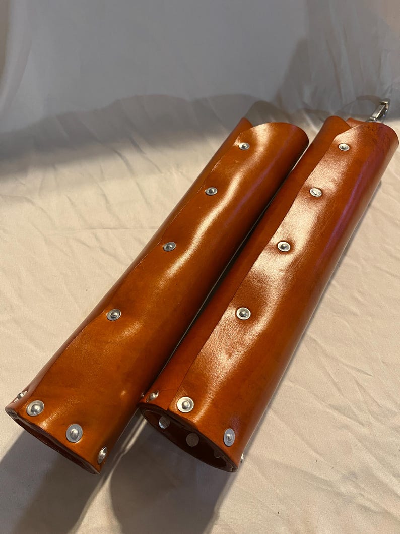 May include: Two brown leather arm guards with silver rivets. The cylindrical guards are open on one side and have a metal clasp at the top. The leather has a glossy finish.