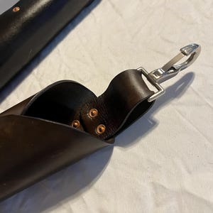 Puede incluir: Primer plano de una correa de cuero marr&oacute;n oscuro con un cierre de metal plateado. La correa est&aacute; asegurada con remaches de cobre. Otra pieza de cuero es visible en el fondo, tambi&eacute;n con remaches de cobre.