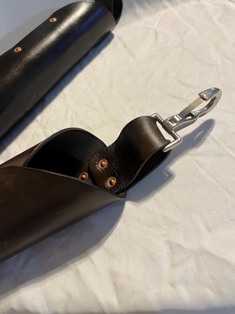 Puede incluir: Primer plano de una correa de cuero marr&oacute;n oscuro con un cierre de metal plateado. La correa est&aacute; asegurada con remaches de cobre. Otra pieza de cuero es visible en el fondo, tambi&eacute;n con remaches de cobre.