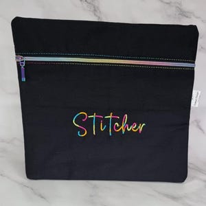 May include: A black zipped pouch featuring a rainbow-coloured zip and matching rainbow-coloured embroidery that reads "Stitcher" in a cursive font. The pouch has a rectangular shape and includes a small white fabric tag on the right side.