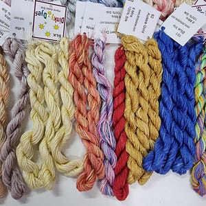 May include: A collection of hand-dyed silk embroidery threads in various colours including yellow, red, blue, and purple. Each skein is twisted and attached to a white paper tag labelled with the brand Dinky Dyes, colour name, and 8 metres length.