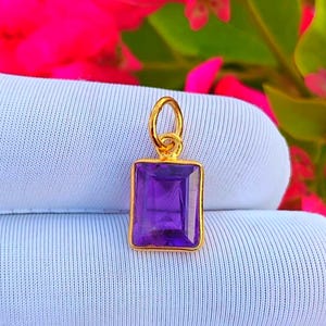 May include: A gold pendant featuring a rectangular, deep purple gemstone. The pendant has a simple design, with a gold loop for a chain. The background is a soft blue fabric and vibrant pink flowers.