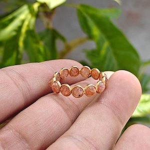 May include: A gold-colored ring featuring a series of round, orange-toned gemstones. The ring is held in a hand, with a blurred green background of foliage. The gemstones are set closely together, creating a continuous band of color.