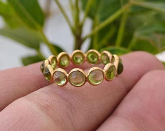 Könnte beinhalten: Ein goldener Ring mit einer Reihe von runden, hellgrünen Edelsteinen. Das Band des Rings hat einen warmen Goldton, und die Edelsteine sind gleichmäßig um das Band angeordnet. Der Ring wird auf einem Finger gezeigt, mit einem verschwommenen grünen Hintergrund.