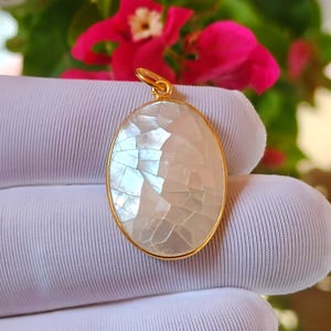May include: An oval pendant featuring a mosaic of iridescent white mother-of-pearl pieces set within a thin gold-toned metal frame. The pendant includes a small gold-toned circular bail at the top for attachment to a chain or cord.