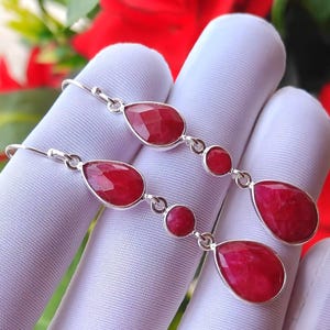 May include: A pair of drop earrings featuring three faceted, teardrop-shaped red gemstones set in silver metal. Each earring consists of a small round red stone positioned between two larger teardrop-shaped red stones, all linked by silver wire.