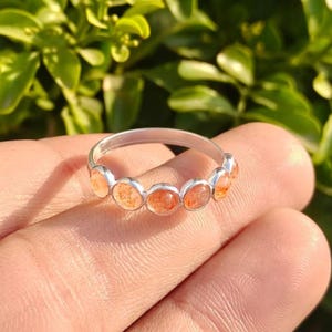 Op de afbeelding: Een zilveren ring met een reeks oranje edelstenen. De edelstenen zijn rond en dicht op elkaar geplaatst, waardoor een doorlopende band van kleur ontstaat. De ring wordt getoond tegen een vage groene achtergrond.
