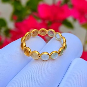 Könnte beinhalten: Ein goldener Ring mit einer Reihe kleiner, runder, in Zargenfassung gefasster Edelsteine in abwechselnden Farbtönen von blassem Gelb und Hellblau. Der Ring hat ein durchgehendes kreisförmiges Design mit polierten Metallfassungen für jeden Stein.