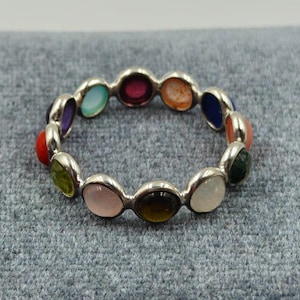 Puede incluir: Un anillo de plata con una serie de piedras preciosas ovaladas de colores. Las piedras preciosas incluyen rojo, naranja, amarillo, verde, azul, morado y rosa. El anillo está sobre un fondo gris.