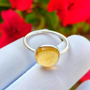 Könnte beinhalten: Ein silberner Ring mit einem großen, quadratisch geschliffenen, gelben Edelstein. Der Ring ist vor einem weißen Stoffhintergrund mit verschwommenen roten Blumen im Hintergrund platziert.