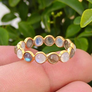 May include: A gold ring featuring a series of round, iridescent gemstones. The gemstones are set closely together, creating a continuous band of color. The ring is held against a backdrop of green foliage.