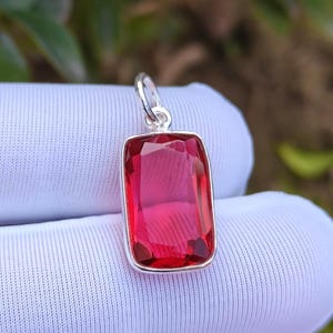 Puede incluir: Un colgante rectangular con una gema roja vibrante engastada en un marco plateado. La gema tiene un corte facetado, captando la luz. El colgante está suspendido de una argolla plateada.