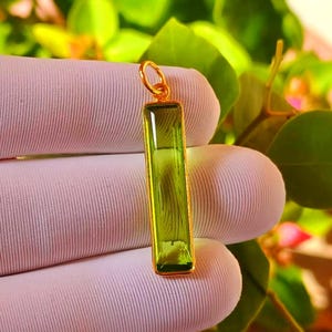 May include: A rectangular, lime green gemstone pendant set in a thin gold-toned metal frame. The pendant features a smooth, faceted surface and a small circular gold-toned bail at the top for attachment to a chain or cord, presented vertically.