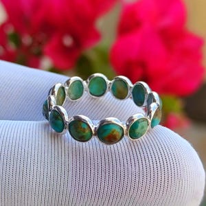 May include: A silver ring set with oval turquoise stones. The turquoise stones display a mix of blue and brown tones. The ring is displayed against a white background, with blurred pink flowers in the background.