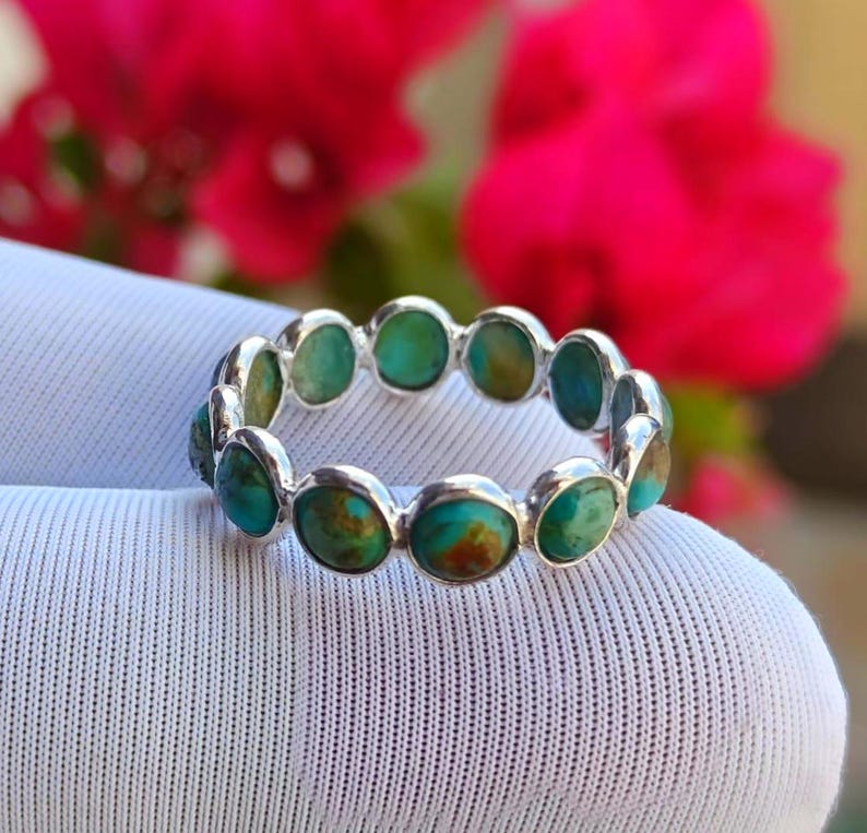 May include: A silver ring set with oval turquoise stones. The turquoise stones display a mix of blue and brown tones. The ring is displayed against a white background, with blurred pink flowers in the background.