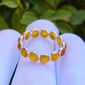 Op de afbeelding: Een zilveren ring met een reeks ronde, amberkleurige edelstenen. De edelstenen zijn dicht op elkaar geplaatst, waardoor een doorlopende band van kleur ontstaat. De ring wordt getoond tegen een witte achtergrond met groen gebladerte op de achtergrond.