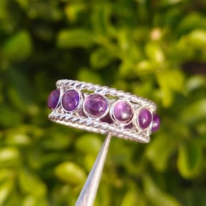 May include: A silver ring with a braided design, featuring six round, deep purple gemstones. The ring is held by a silver tool against a blurred green background, showcasing the jewelry's intricate details and color.