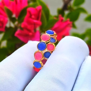 Pode incluir: Um anel de ouro com pedras circulares rosa e azuis alternadas. O anel é segurado contra um fundo branco, com flores rosa e folhas verdes ao fundo.