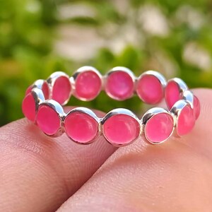 Peut inclure: Une bague en argent avec une bande continue de pierres précieuses ovales rose vif. Les pierres sont serties de près, créant un cercle complet de couleur. La bague est présentée sur un fond vert flou.