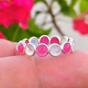 Pode incluir: Um anel de prata com pedras preciosas rosa e brancas alternadas. O anel apresenta uma série de pedras redondas e polidas embutidas em uma faixa contínua. As pedras preciosas são de um rosa vibrante e um branco translúcido.