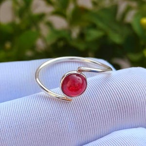 May include: A silver ring with a bright red gemstone. The ring has a slender, curved band that encircles the finger, with the gemstone set in a circular bezel. The red stone is the main feature, providing a striking contrast.