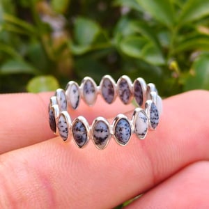 May include: A silver ring with a repeating pattern of oval-shaped stones. The stones have a speckled design in black and grey on a white base. The ring is set against a blurred green backdrop.