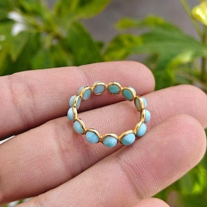May include: A gold ring featuring a series of light blue gemstones. The ring is set in a gold band, with each gemstone individually set. The ring is shown against a background of green foliage.
