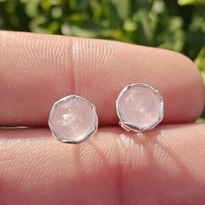 Può includere: Due orecchini a lobo in quarzo rosa con forma rotonda sfaccettata e castone color argento. Gli orecchini sono posti su uno sfondo sfocato, evidenziando le pietre preziose rosa.