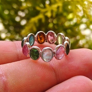 Puede incluir: Un anillo de plata con una serie de piedras preciosas ovaladas en varios colores, incluyendo rosa, verde, azul y naranja. El anillo se muestra en un dedo, con un fondo verde borroso.