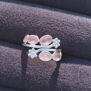 Puede incluir: Un anillo de plata con un diseño único, con múltiples piedras preciosas rosas de forma ovalada. El anillo tiene una banda dividida con detalles florales y está sobre un fondo oscuro y texturizado.