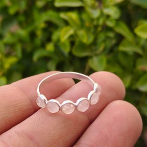 May include: A silver ring featuring a series of pale pink, round gemstones. The ring is set with the gemstones evenly spaced around the band. The ring is shown against a blurred green background.