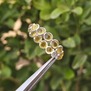 Könnte beinhalten: Ein silberner Ring mit einer Reihe runder, gelb-grüner Edelsteine. Der Ring wird von einer silbernen Pinzette vor einem verschwommenen grünen Hintergrund aus Blättern gehalten. Die Edelsteine sind gleichmäßig angeordnet.