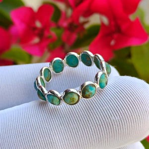 May include: A silver ring with a repeating pattern of oval turquoise stones. The ring is displayed on a white surface, with blurred red flowers in the background. The turquoise stones display a mix of blue and green colours.