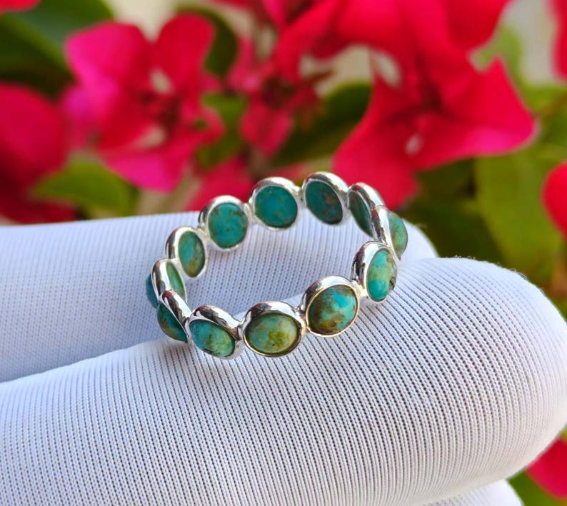 May include: A silver ring with a repeating pattern of oval turquoise stones. The ring is displayed on a white surface, with blurred red flowers in the background. The turquoise stones display a mix of blue and green colours.
