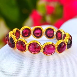 Può includere: Un anello d'oro con una serie di pietre preziose ovali rosso scuro incastonate in castoni individuali. L'anello è esposto su una superficie bianca, con uno sfondo sfocato verde e rosa.