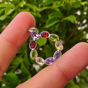 以下が含まれることがあります： カラフルな宝石が並んだ、円形のデザインのシルバーペンダント。宝石は楕円形で、赤、緑、紫、黄色の色合いが含まれています。ペンダントは、ぼやけた緑色の背景に対して、2本の指の間に保持されています。