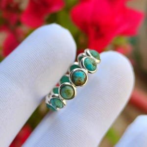 May include: A silver ring with a row of turquoise gemstones. Each round turquoise stone is set in its own bezel, forming a continuous band. The ring is shown against a white background, with red flowers visible in the background.