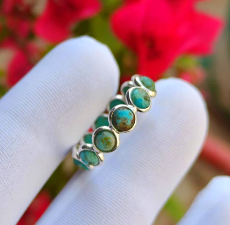 May include: A silver ring with a row of turquoise gemstones. Each round turquoise stone is set in its own bezel, forming a continuous band. The ring is shown against a white background, with red flowers visible in the background.