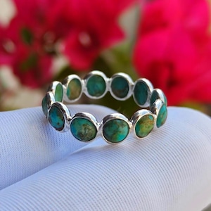 May include: A silver ring set with turquoise stones. The ring has a repeating pattern of round turquoise stones, each set in a silver bezel. The background features blurred pink flowers, with the ring resting on a white surface.